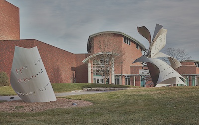 Red brick building with sloped roof in the background, abstract art installation on the lawn that uses large book pages.