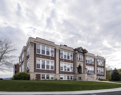 Large three story rectangular brick building with tall windows, stairs leading to the entrance, and two large columns by the door.