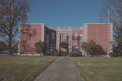 Outside of a large red brick building, which sits on a field of grass and a paved sidewalk leads up to the front door. Trees border the building. 