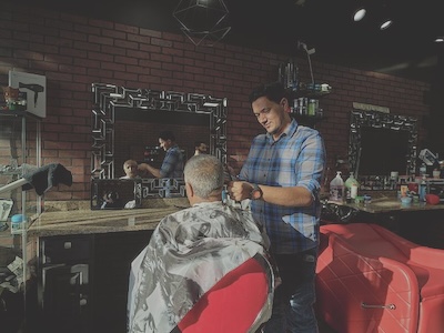 A barber cuts a gray-haired man's hair in front of a mirror with an ornate border.