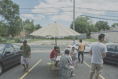 Outdoors in a parking spot between two parked cars four men play cards at a table.